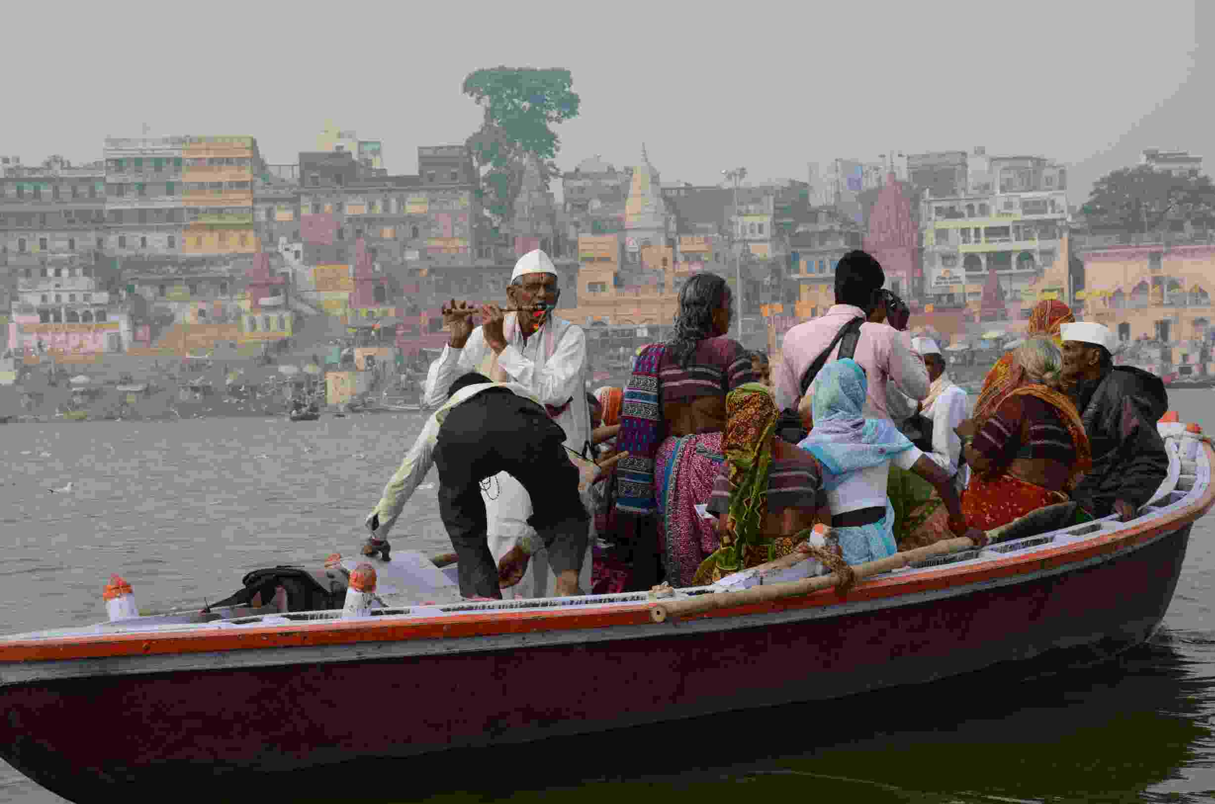 Image shows a man playing a pipe in boat on the River Ganges, amongst a group of brightly dressed people